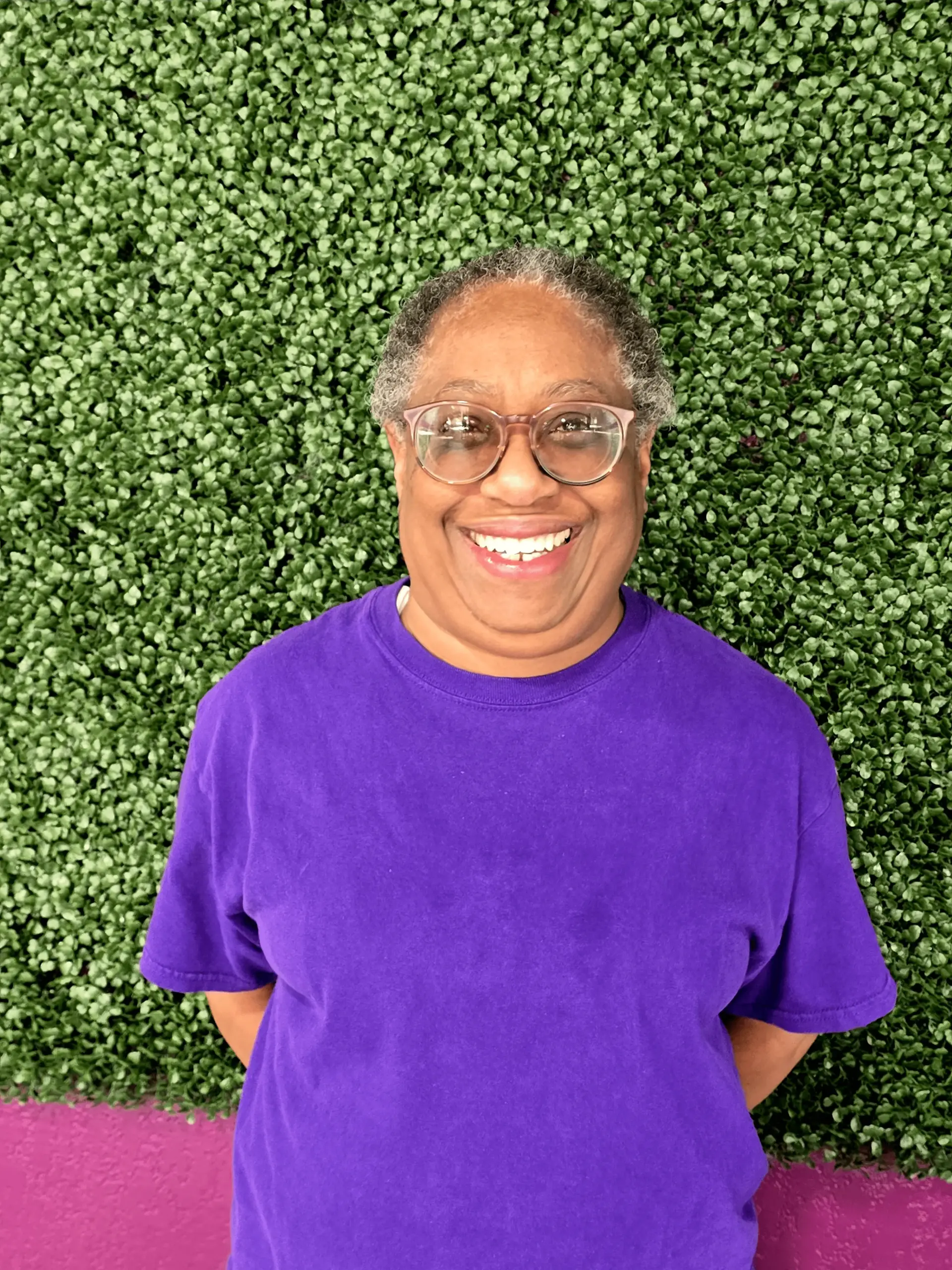A smiling flyer in a purple tee photographed head-on against the studio’s green hedge wall