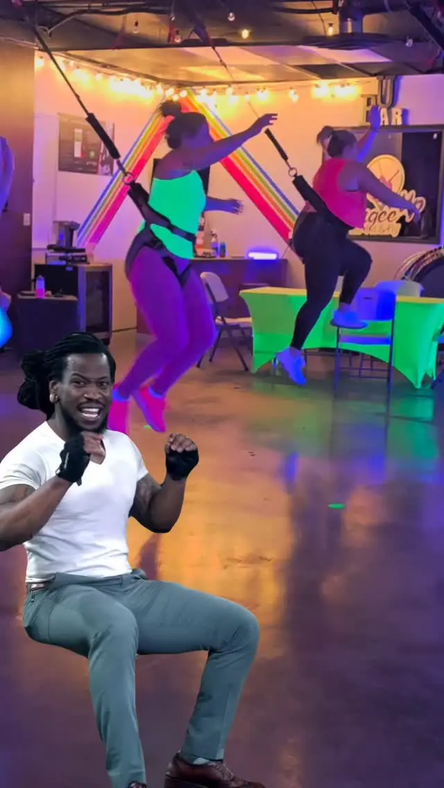 A coach leading a squat on the studio floor while two flyers hang mid-air behind him, neon decorations along the back wall